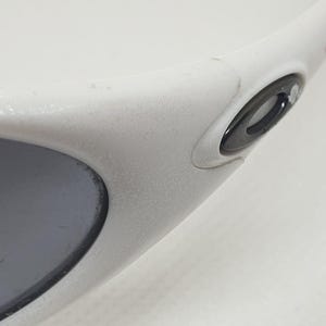 Puede incluir: Primer plano de gafas de sol blancas con lente gris. Las gafas tienen una montura redondeada y un logotipo negro en la patilla. La montura parece ser de material pl&aacute;stico mate.