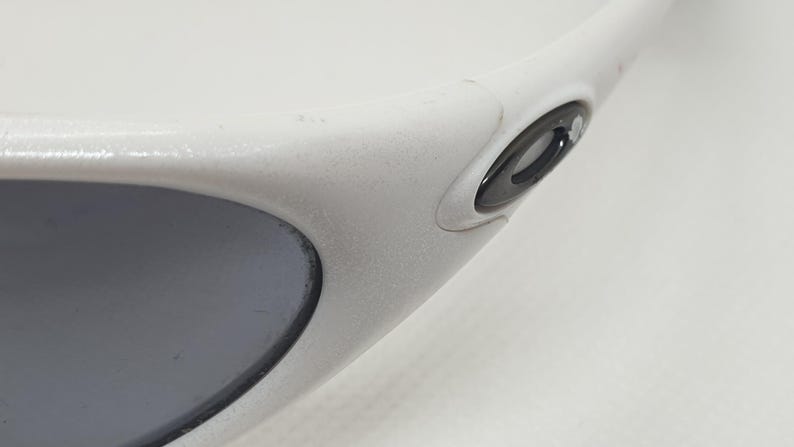 Puede incluir: Primer plano de gafas de sol blancas con lente gris. Las gafas tienen una montura redondeada y un logotipo negro en la patilla. La montura parece ser de material pl&aacute;stico mate.
