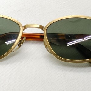 May include: Gold-framed sunglasses with green lenses. The frames have a rounded shape and a bridge connecting the lenses. The temples are a tortoiseshell pattern. The sunglasses are designed for eye protection.