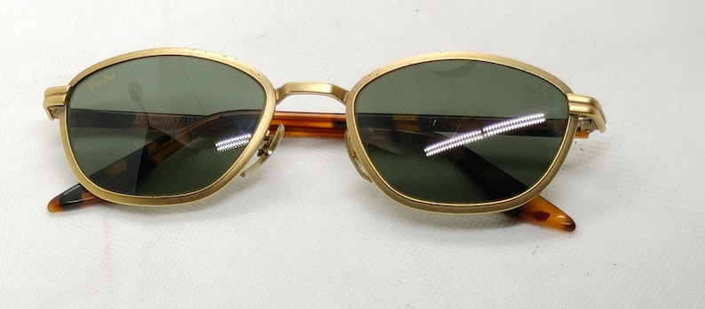 May include: Gold-framed sunglasses with green lenses. The frames have a rounded shape and a bridge connecting the lenses. The temples are a tortoiseshell pattern. The sunglasses are designed for eye protection.