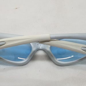 K&ouml;nnte beinhalten: Zusammengeklappte hellgraue Sonnenbrille mit blauen Gl&auml;sern. Die B&uuml;gel sind hellgrau und wei&szlig;. Die Gl&auml;ser sind hellblau. Die Sonnenbrille liegt auf einer wei&szlig;en Oberfl&auml;che.
