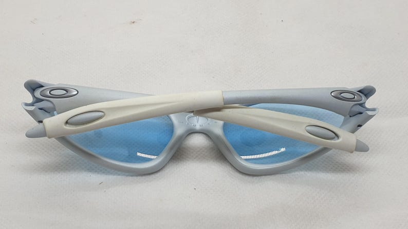 K&ouml;nnte beinhalten: Zusammengeklappte hellgraue Sonnenbrille mit blauen Gl&auml;sern. Die B&uuml;gel sind hellgrau und wei&szlig;. Die Gl&auml;ser sind hellblau. Die Sonnenbrille liegt auf einer wei&szlig;en Oberfl&auml;che.