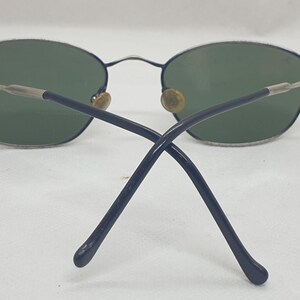 Puede incluir: Gafas de sol ovaladas con lentes verdes. Las monturas son plateadas y azul marino, con un fino puente y varillas met&aacute;licas. Las varillas se cruzan en la parte trasera.