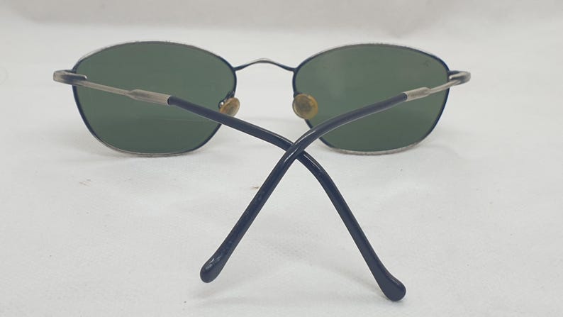 Puede incluir: Gafas de sol ovaladas con lentes verdes. Las monturas son plateadas y azul marino, con un fino puente y varillas met&aacute;licas. Las varillas se cruzan en la parte trasera.