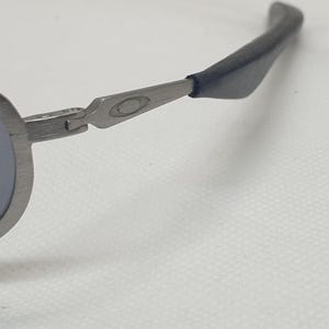 K&ouml;nnte beinhalten: Nahaufnahme einer silberfarbenen Sonnenbrille mit blauen Gl&auml;sern. Die Sonnenbrille hat ein schlankes, modernes Design mit einem geschwungenen Rahmen und dunkelgrauen B&uuml;geln. Das Oakley-Logo ist auf dem B&uuml;gel sichtbar.