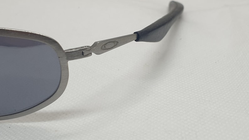 K&ouml;nnte beinhalten: Nahaufnahme einer silberfarbenen Sonnenbrille mit blauen Gl&auml;sern. Die Sonnenbrille hat ein schlankes, modernes Design mit einem geschwungenen Rahmen und dunkelgrauen B&uuml;geln. Das Oakley-Logo ist auf dem B&uuml;gel sichtbar.