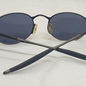 May include: Black oval sunglasses with dark lenses. The sunglasses have a thin, black metal frame and temples. The nose pads are gold-toned. The arms are crossed, showing the details of the design.