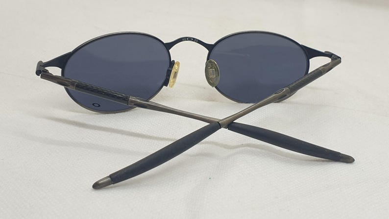 May include: Black oval sunglasses with dark lenses. The sunglasses have a thin, black metal frame and temples. The nose pads are gold-toned. The arms are crossed, showing the details of the design.