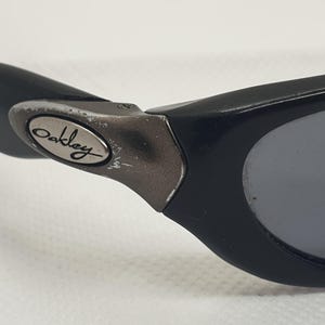 May include: Close-up of black Oakley sunglasses. The sunglasses have a black frame with grey lenses. The Oakley logo is visible on the temple arm. The sunglasses are designed for sports and outdoor activities.