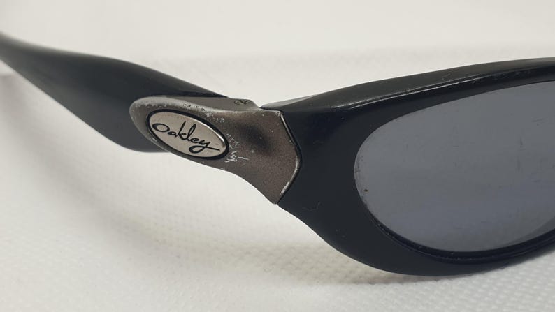 May include: Close-up of black Oakley sunglasses. The sunglasses have a black frame with grey lenses. The Oakley logo is visible on the temple arm. The sunglasses are designed for sports and outdoor activities.