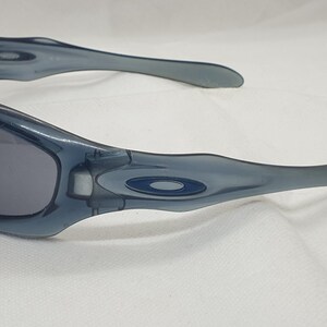 May include: A pair of blue-grey sunglasses with dark lenses. The sunglasses have a curved frame and temples, with a small logo on the side. The lenses are a dark grey color, and the frame is a translucent blue-grey.