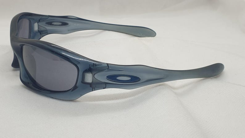 May include: A pair of blue-grey sunglasses with dark lenses. The sunglasses have a curved frame and temples, with a small logo on the side. The lenses are a dark grey color, and the frame is a translucent blue-grey.