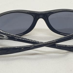 Puede incluir: Gafas de sol negras con lentes grises. Las gafas de sol tienen un acabado brillante y un dise&ntilde;o curvo. Las patillas de las gafas est&aacute;n cruzadas, mostrando el logotipo de la marca. Las lentes son de color gris oscuro.
