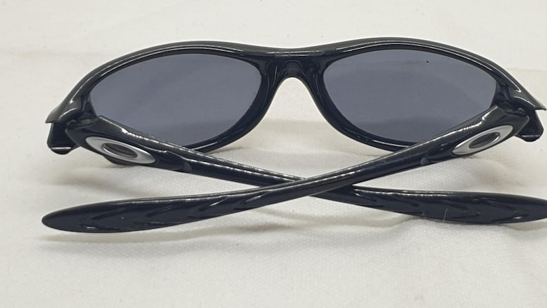 Puede incluir: Gafas de sol negras con lentes grises. Las gafas de sol tienen un acabado brillante y un dise&ntilde;o curvo. Las patillas de las gafas est&aacute;n cruzadas, mostrando el logotipo de la marca. Las lentes son de color gris oscuro.