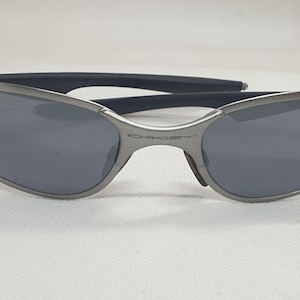 K&ouml;nnte beinhalten: Graue Oakley-Sonnenbrille mit dunkelgrauen Gl&auml;sern. Die Sonnenbrille hat ein schlankes, geschwungenes Design mit einem Metallrahmen und schwarzen B&uuml;geln. Das Oakley-Logo ist sichtbar.