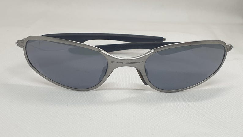 K&ouml;nnte beinhalten: Graue Oakley-Sonnenbrille mit dunkelgrauen Gl&auml;sern. Die Sonnenbrille hat ein schlankes, geschwungenes Design mit einem Metallrahmen und schwarzen B&uuml;geln. Das Oakley-Logo ist sichtbar.