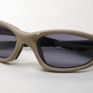 Puede incluir: Gafas de sol de color beige con lentes oscuras. Las gafas tienen una montura curvada y un dise&ntilde;o &uacute;nico en las patillas. Las lentes son de color gris oscuro y la montura es de color beige liso. Las gafas est&aacute;n sobre un fondo blanco.