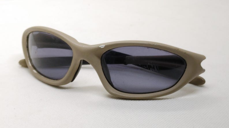 Puede incluir: Gafas de sol de color beige con lentes oscuras. Las gafas tienen una montura curvada y un dise&ntilde;o &uacute;nico en las patillas. Las lentes son de color gris oscuro y la montura es de color beige liso. Las gafas est&aacute;n sobre un fondo blanco.