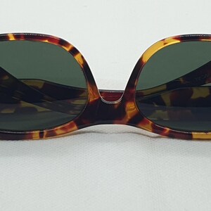 Puede incluir: Gafas de sol carey con lentes verde oscuro. Las monturas tienen una forma cl&aacute;sica y redondeada con un patr&oacute;n marr&oacute;n y amarillo. Las gafas de sol est&aacute;n sobre un fondo blanco.
