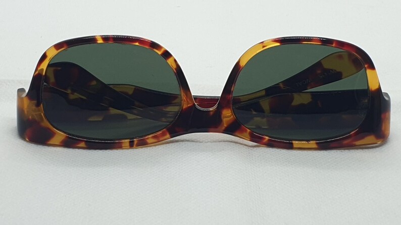Puede incluir: Gafas de sol carey con lentes verde oscuro. Las monturas tienen una forma cl&aacute;sica y redondeada con un patr&oacute;n marr&oacute;n y amarillo. Las gafas de sol est&aacute;n sobre un fondo blanco.