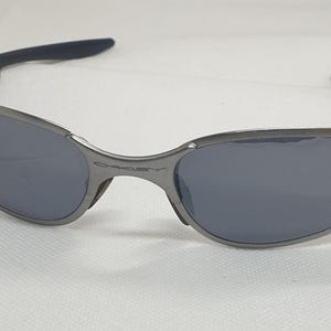 K&ouml;nnte beinhalten: Graue Sonnenbrille mit dunklen Gl&auml;sern. Die Rahmen sind metallisch grau und die B&uuml;gel haben schwarze, gummierte Griffe. Die Gl&auml;ser sind dunkelgrau und sorgen f&uuml;r einen eleganten, modernen Look. Die Sonnenbrille ist f&uuml;r den Augenschutz konzipiert.