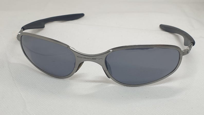K&ouml;nnte beinhalten: Graue Sonnenbrille mit dunklen Gl&auml;sern. Die Rahmen sind metallisch grau und die B&uuml;gel haben schwarze, gummierte Griffe. Die Gl&auml;ser sind dunkelgrau und sorgen f&uuml;r einen eleganten, modernen Look. Die Sonnenbrille ist f&uuml;r den Augenschutz konzipiert.