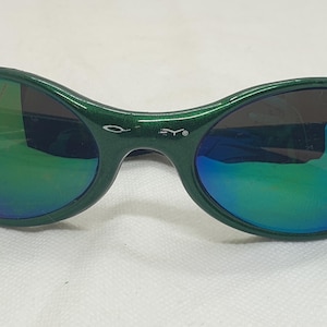 May include: Green oval-shaped sunglasses with iridescent lenses. The frames are a solid green color, and the lenses reflect a rainbow of colors. The sunglasses have a small logo on the bridge.