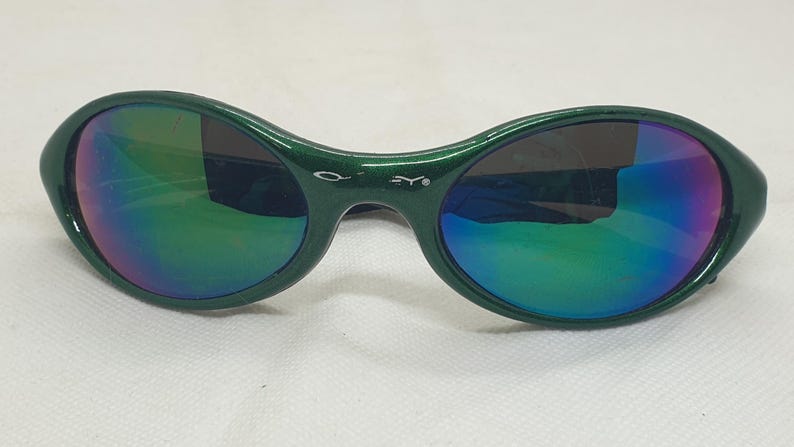 May include: Green oval-shaped sunglasses with iridescent lenses. The frames are a solid green color, and the lenses reflect a rainbow of colors. The sunglasses have a small logo on the bridge.