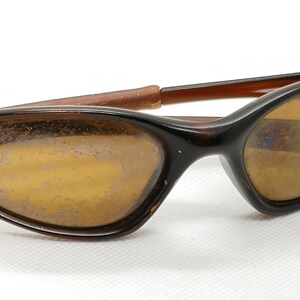 Puede incluir: Gafas de sol marrones con montura curvada y lentes tintadas en &aacute;mbar. Las gafas de sol tienen un dise&ntilde;o retro, con una forma elegante y aerodin&aacute;mica. Las patillas son de color marr&oacute;n claro y las lentes son ligeramente reflectantes.