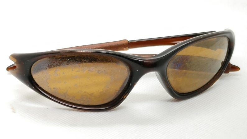 Puede incluir: Gafas de sol marrones con montura curvada y lentes tintadas en &aacute;mbar. Las gafas de sol tienen un dise&ntilde;o retro, con una forma elegante y aerodin&aacute;mica. Las patillas son de color marr&oacute;n claro y las lentes son ligeramente reflectantes.