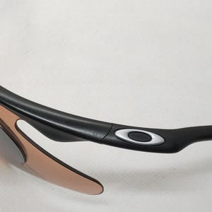 May include: Close-up of black sunglasses with brown-tinted lenses. The sunglasses have a sleek, modern design with a black frame and the Oakley logo in silver on the temple.