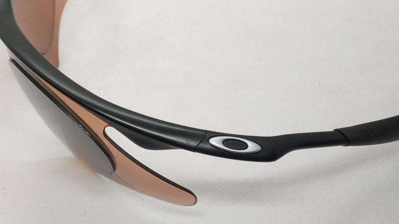 May include: Close-up of black sunglasses with brown-tinted lenses. The sunglasses have a sleek, modern design with a black frame and the Oakley logo in silver on the temple.