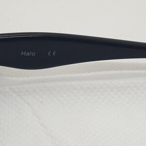 May include: Close-up of a black eyeglass arm with the word "Halo" and the letters "CE" printed on it. The arm is attached to the frame with two small screws.