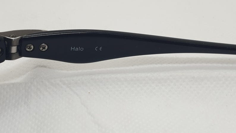 May include: Close-up of a black eyeglass arm with the word "Halo" and the letters "CE" printed on it. The arm is attached to the frame with two small screws.