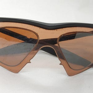 May include: Unusual sunglasses featuring a black frame and brown lenses. The lenses have a distinctive shape, with a pointed edge on the bottom. The sunglasses have a retro or vintage aesthetic.