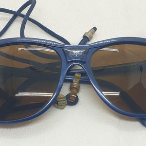 May include: Dark blue sunglasses with brown tinted lenses and a navy blue cord strap. The bridge of the frame features a small emblem with the text KVARNER. The design includes a thick plastic frame and a cord that loops behind the lenses.