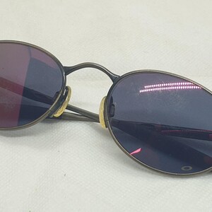 May include: Oval-shaped sunglasses with a dark metal frame and tinted lenses. The lenses have a reflective quality, showing shades of purple and red. The arms of the glasses are thin and extend to the temples.