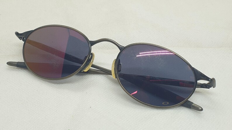 May include: Oval-shaped sunglasses with a dark metal frame and tinted lenses. The lenses have a reflective quality, showing shades of purple and red. The arms of the glasses are thin and extend to the temples.