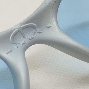 K&ouml;nnte beinhalten: Nahaufnahme einer hellgrauen Sonnenbrille mit blauen Gl&auml;sern. Der Rahmen hat ein Logo auf der Br&uuml;cke. Die Gl&auml;ser sind hellblau. Die Sonnenbrille besteht aus einem matten Material.