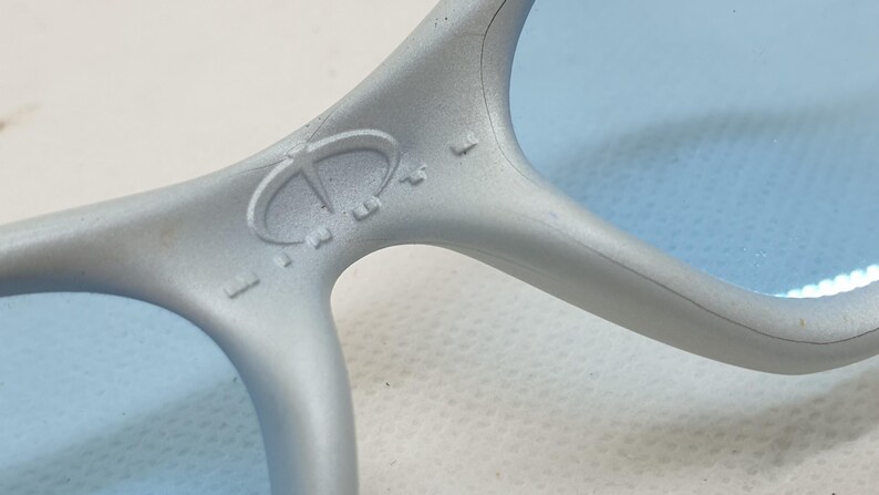 K&ouml;nnte beinhalten: Nahaufnahme einer hellgrauen Sonnenbrille mit blauen Gl&auml;sern. Der Rahmen hat ein Logo auf der Br&uuml;cke. Die Gl&auml;ser sind hellblau. Die Sonnenbrille besteht aus einem matten Material.