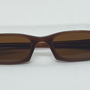 Pode incluir: &Oacute;culos de sol castanhos com lentes castanhas. As arma&ccedil;&otilde;es retangulares t&ecirc;m uma parte superior ligeiramente curva e est&atilde;o sobre um fundo branco. As hastes s&atilde;o azuis escuras.