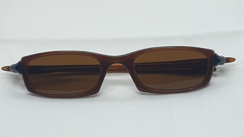 Pode incluir: &Oacute;culos de sol castanhos com lentes castanhas. As arma&ccedil;&otilde;es retangulares t&ecirc;m uma parte superior ligeiramente curva e est&atilde;o sobre um fundo branco. As hastes s&atilde;o azuis escuras.