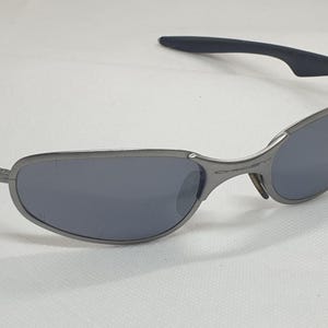 K&ouml;nnte beinhalten: Graue Sonnenbrille mit dunklen Gl&auml;sern. Die Fassungen sind metallisch grau, und die B&uuml;gel haben schwarze Gummigriffe. Die Gl&auml;ser sind dunkelgrau und bieten Sonnenschutz.