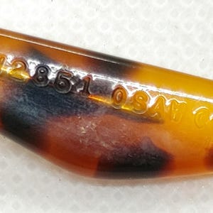 May include: Close-up of a vintage, tortoiseshell-patterned object with a pointed end. The object has a glossy finish and features the text "H28510SAT CO" etched into its surface. The color palette includes shades of amber, brown, and black.