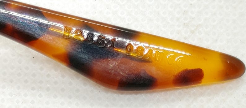 May include: Close-up of a vintage, tortoiseshell-patterned object with a pointed end. The object has a glossy finish and features the text "H28510SAT CO" etched into its surface. The color palette includes shades of amber, brown, and black.