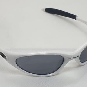 Puede incluir: Gafas de sol blancas con lentes grises y puntas de patillas negras. Las gafas de sol tienen un dise&ntilde;o de montura curvada y est&aacute;n sobre un fondo blanco. Las lentes son de forma ovalada.