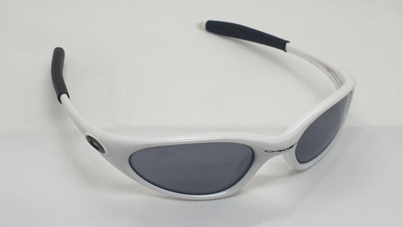 Puede incluir: Gafas de sol blancas con lentes grises y puntas de patillas negras. Las gafas de sol tienen un dise&ntilde;o de montura curvada y est&aacute;n sobre un fondo blanco. Las lentes son de forma ovalada.