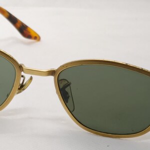 May include: Gold-framed sunglasses with green lenses. The frames have a classic oval shape and are accented with tortoise-shell patterned arms. The sunglasses are designed for eye protection and fashion.