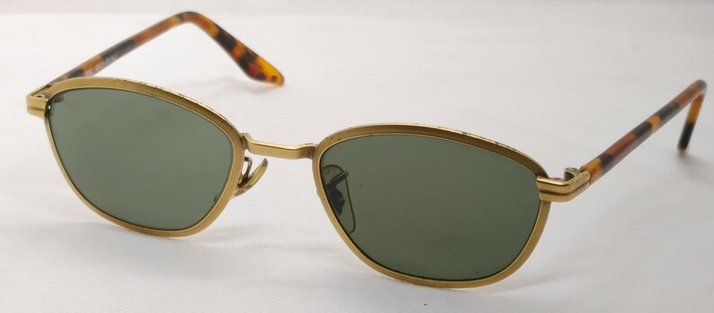 May include: Gold-framed sunglasses with green lenses. The frames have a classic oval shape and are accented with tortoise-shell patterned arms. The sunglasses are designed for eye protection and fashion.