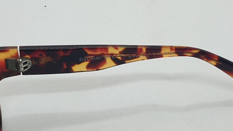 Puede incluir: Primer plano de la patilla de unas gafas con estampado de carey. El texto "B & RAY BAN" es visible en la patilla. Las gafas tienen una bisagra plateada y un dise&ntilde;o curvado.
